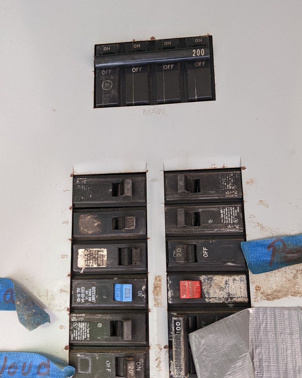 An electrical panel with the main and branch breakers switched off, several of them covered with tape and marked in pen.